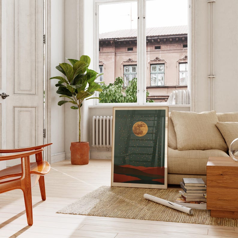 May include: A framed art print featuring a full moon over a landscape with a ladder. The artwork has a dark green background with a yellow moon and red and brown hills. The print is displayed in a bright, airy room with a wooden chair and a large potted plant.
