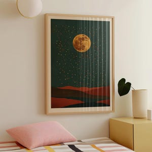 May include: A framed art print depicting a night landscape. The artwork features a large, textured orange moon against a dark green sky filled with stars. Below the sky are red and brown hills. The print is in a light wood frame.