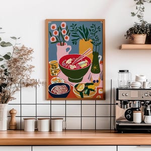 May include: A framed art print depicting a colorful still life of a meal, including a bowl of soup with chopsticks, a side of noodles, and a plate with lemon slices. The artwork is set against a blue background with pink and yellow accents.
