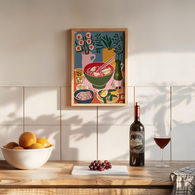 May include: A framed art print depicts a colorful illustration of a bowl of ramen with chopsticks, alongside other dishes. A bottle of wine, a glass of red wine, and a bowl of oranges are on a wooden surface. A bunch of grapes sits on a white cutting board.