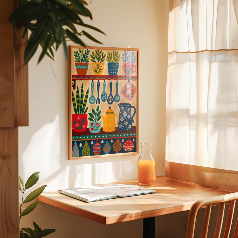 May include: A framed art print featuring a colorful kitchen scene with potted plants, utensils, and a blue floral pitcher. The artwork is displayed on a wall above a wooden table with a glass pitcher of orange juice and an open book.