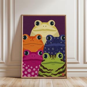 May include: A framed art print featuring five stylized frogs in a vertical arrangement. The top frog is pale yellow, with an orange frog and a dark blue star-patterned frog below it. At the bottom are a magenta polka-dot frog and a green striped frog.