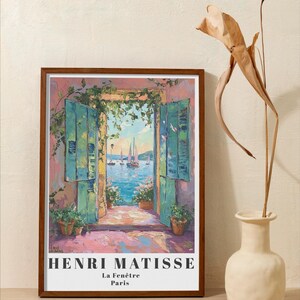 Peut inclure: Une impression encadr&eacute;e d'une peinture d'Henri Matisse intitul&eacute;e "La Fen&ecirc;tre" repr&eacute;sentant une vue d'un paysage marin &agrave; travers une porte ouverte avec des volets verts. La peinture est sur un fond rose.
