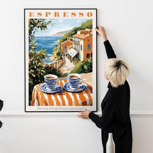 May include: A framed poster with a colorful illustration of a seaside town with two cups of coffee on a striped table. The poster has the word "ESPRESSO" in large letters at the top.