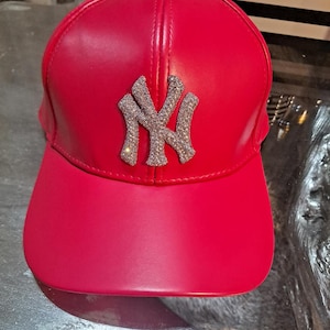 May include: A vibrant red baseball cap featuring a sparkling silver "NY" logo. The cap has a curved brim and a structured design. This stylish accessory is perfect for adding a pop of color to any outfit.