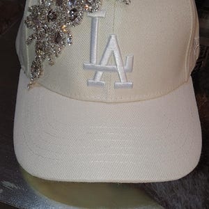 May include: Cream-colored baseball cap with the letters "LA" embroidered on the front. The cap is embellished with a sparkling, ornate crystal design on the side.