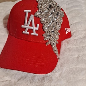 May include: A red baseball cap with the white letters "LA" embroidered on the front. The cap is embellished with a large, sparkling crystal design on the side. The New Era logo is visible on the side.
