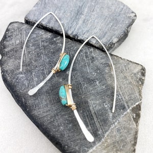 May include: A pair of silver earrings with turquoise stones wrapped in gold wire. The earrings are hanging from a gray stone background.