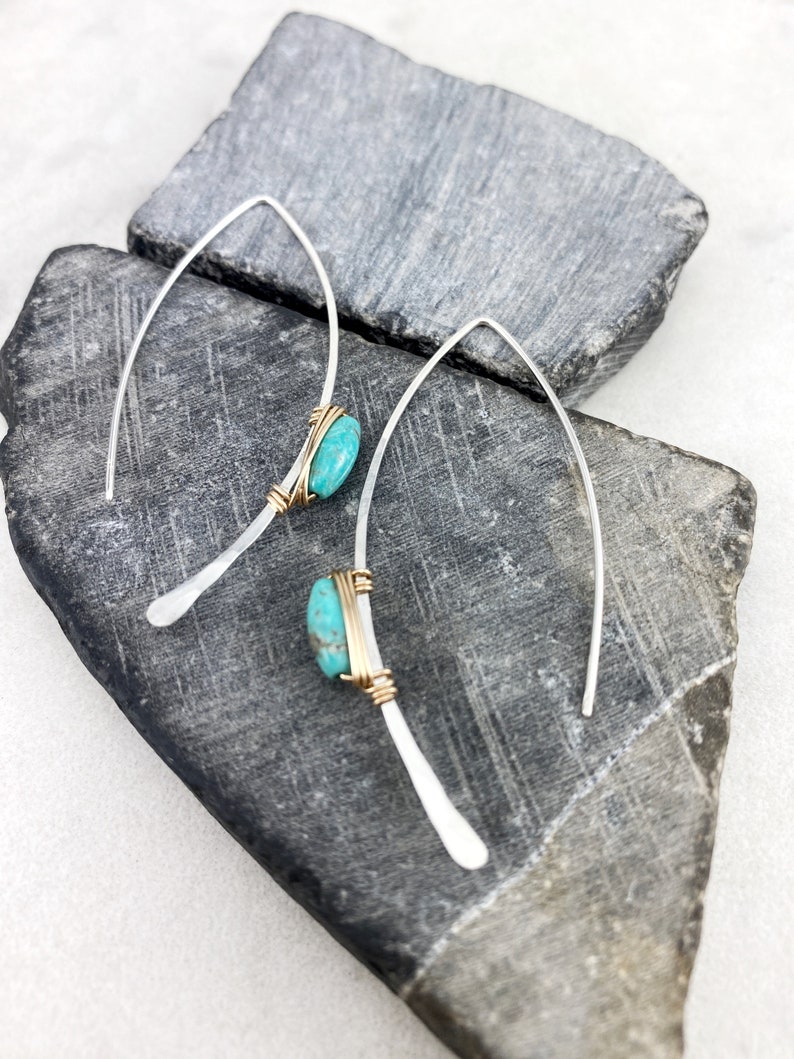 May include: A pair of silver earrings with turquoise stones wrapped in gold wire. The earrings are hanging from a gray stone background.