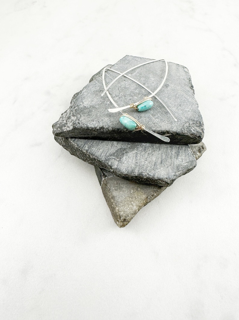 May include: A pair of silver earrings with turquoise stones set in gold wire. The earrings are displayed on a stack of gray stones.