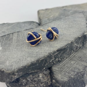 May include: A pair of gold wire wrapped lapis lazuli earrings. The earrings are a dark blue color and are set on a gray stone background.