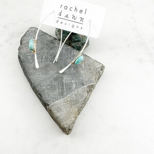 May include: A pair of silver hoop earrings with a turquoise stone wrapped in gold wire. The earrings are displayed on a gray rock with a white card that reads "rachel dawn designs".