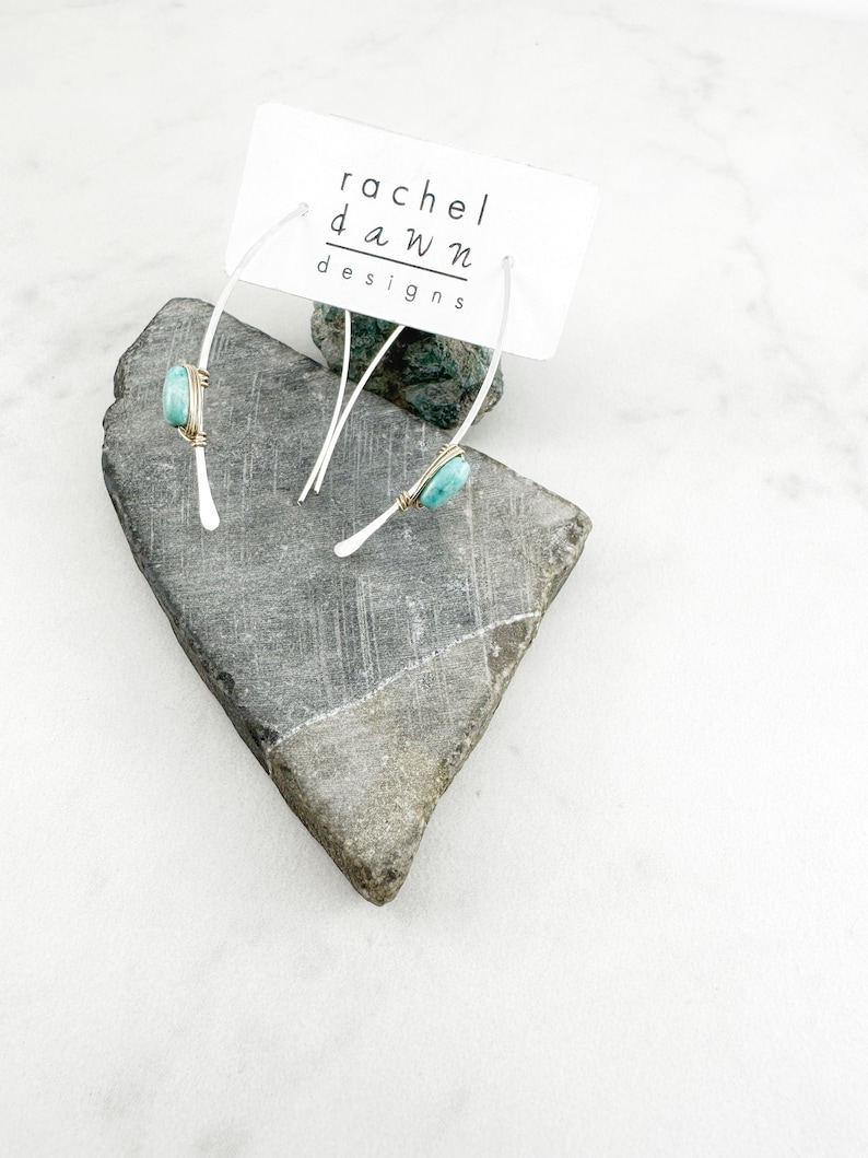 May include: A pair of silver hoop earrings with a turquoise stone wrapped in gold wire. The earrings are displayed on a gray rock with a white card that reads "rachel dawn designs".