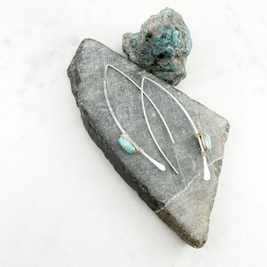 May include: A pair of silver earrings with turquoise stones and gold wire accents. The earrings are displayed on a gray stone.