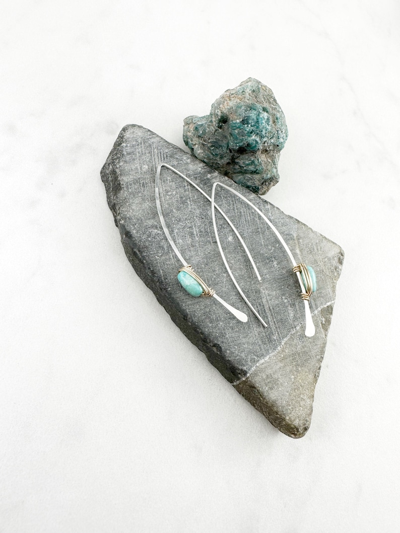 May include: A pair of silver earrings with turquoise stones and gold wire accents. The earrings are displayed on a gray stone.