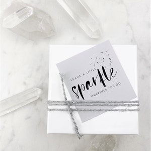May include: A white gift box with a gray tag tied with twine. The tag has the text "Leave a little sparkle wherever you go" in black lettering.