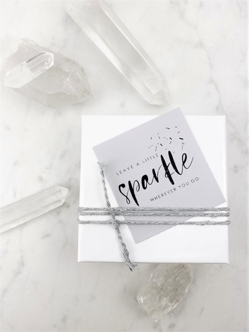 May include: A white gift box with a gray tag tied with twine. The tag has the text "Leave a little sparkle wherever you go" in black lettering.