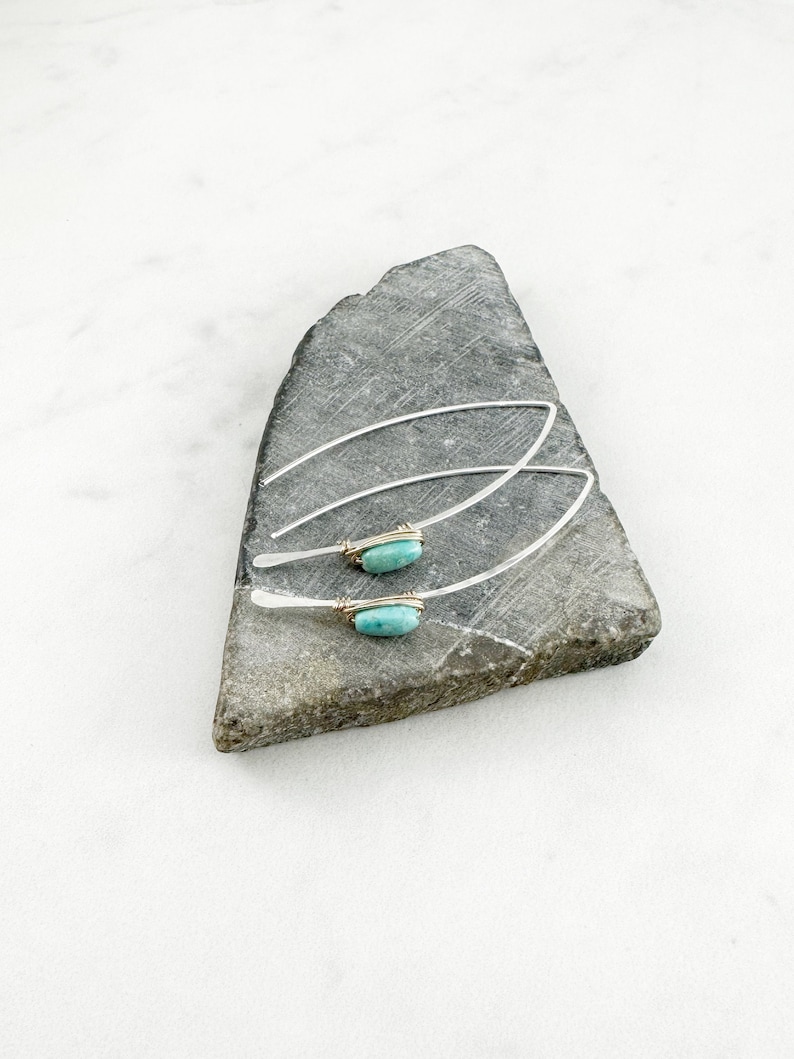 May include: A pair of silver earrings with turquoise stones set in gold wire. The earrings are displayed on a gray stone.