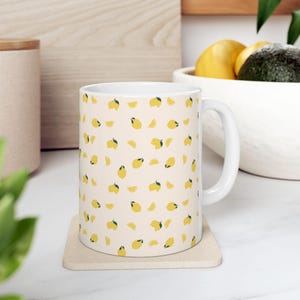 Pode incluir: Caneca de cerâmica branca com um padrão de limões amarelos e fatias de limão em um fundo creme.