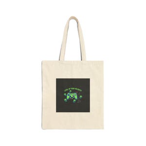 Puede incluir: Bolso tote de color crema con un diseño gráfico cuadrado negro. El diseño presenta un mando de videojuegos verde y azul, cajas de regalo y el texto "LEVEL UP YOUR HOLIDAYS!" en verde. El bolso tiene un diseño sencillo con un asa larga.