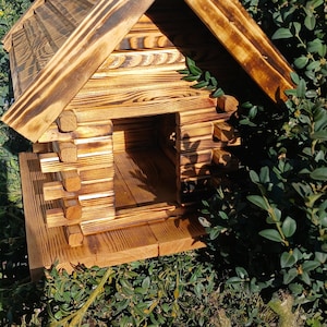 Può includere: Una casetta per uccelli in legno fatta a mano con un design rustico a capanna. La casetta per uccelli presenta un tetto spiovente, un piccolo camino e un ingresso. I toni naturali del legno e lo stile capanna creano un'estetica affascinante. La casetta per uccelli è circondata da verde.