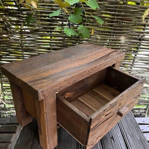 Op de afbeelding: Een kleine, rechthoekige houten kruk met een enkele uitschuifbare lade met een kleine houten knop. Het meubel is gemaakt van donker, verweerd hout met een zichtbare nerf en heeft een rustieke, onafgewerkte textuur over het hele frame.