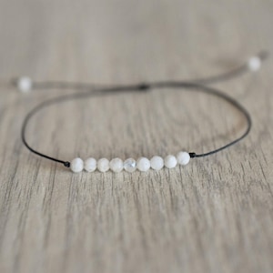 Può includere: Un delicato braccialetto con una serie di piccole perline bianche e rotonde infilate su un sottile cordoncino nero. Il braccialetto ha un design regolabile con una chiusura a nodo scorrevole. Le perline sono distanziate in modo uniforme, creando un'estetica minimalista ed elegante.