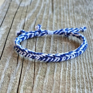 May include: A blue and white braided bracelet. The bracelet is made of woven cord in a chevron pattern. The bracelet is lying on a weathered wooden surface. The bracelet is adjustable.