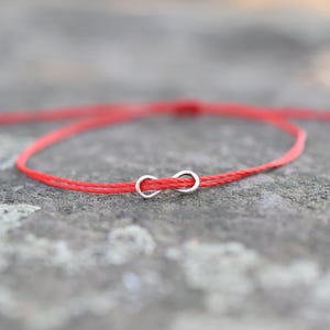 Può includere: Un braccialetto con filo rosso e ciondolo a infinito argentato. Il braccialetto è semplice e delicato, con il simbolo dell'infinito centrato sul filo rosso. Lo sfondo è una superficie grigia strutturata.