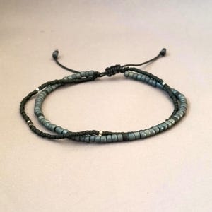 Op de afbeelding: Een dubbelstrengs armband met één streng kleine matzwarte kralen en één streng metallic blauwgrijze kralen. Beide strengen zijn voorzien van kleine zilverkleurige tussenkralen en verbonden door een zwart koord met een verstelbare schuifknoop.
