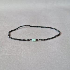 Könnte beinhalten: Ein zartes Armband aus schwarzen Perlen mit einem kleinen, hellgrünen Stein in der Mitte. Das Armband ist auf einem dünnen, flexiblen Band aufgefädelt und wird vor einem neutralen, grauen Hintergrund präsentiert. Dieses minimalistische Schmuckstück ist für den Alltag geeignet.