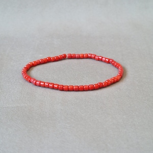 Op de afbeelding: Een rode koraal kralen armband. De armband is gemaakt van kleine, vierkante kralen die aan elkaar geregen zijn op een dun koord.