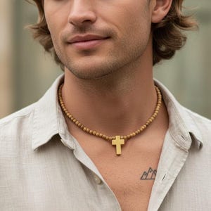 May include: A necklace featuring a series of small, round, tan-colored beads strung together, centered with a smooth, solid gold-toned cross pendant. The cross has a simple, clean-lined design and hangs vertically from the beaded chain.