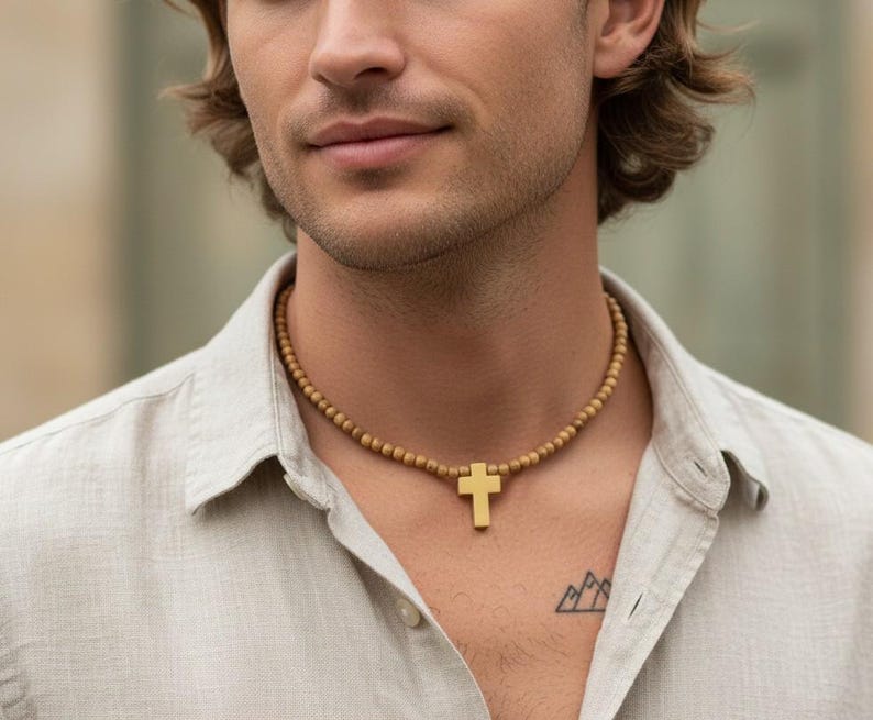 May include: A necklace featuring a series of small, round, tan-colored beads strung together, centered with a smooth, solid gold-toned cross pendant. The cross has a simple, clean-lined design and hangs vertically from the beaded chain.