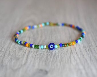 Può includere: Un braccialetto di perline colorate con un ciondolo a forma di occhio blu. Il braccialetto è realizzato con piccole perline rotonde in tonalità di blu, verde, arancione e bianco.
