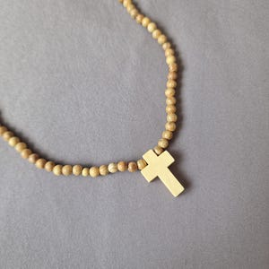 May include: A necklace featuring a series of small, round, light brown wooden beads strung together, accented with a single, smooth, light-colored wooden cross pendant. The text "Azul Sereno Jewelry" is printed in the bottom left corner of the image.