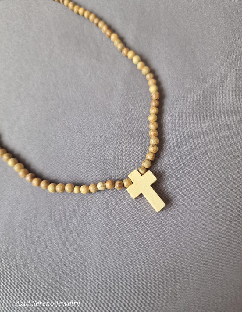 May include: A necklace featuring a series of small, round, light brown wooden beads strung together, accented with a single, smooth, light-colored wooden cross pendant. The text "Azul Sereno Jewelry" is printed in the bottom left corner of the image.