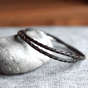Op de afbeelding: Twee gevlochten zwarte leren armbanden, liggend op een grijze steen.