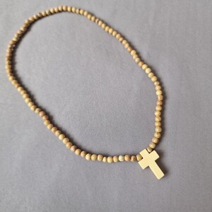 May include: A necklace featuring a continuous strand of small, round, light brown wooden beads and a single, smooth, light-colored cross pendant. The text "Azul Sereno Jewelry" is printed in the bottom left corner of the image.