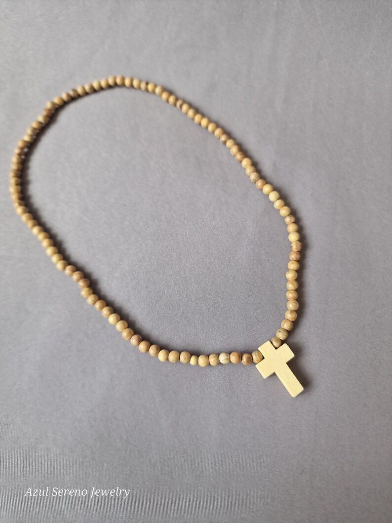 May include: A necklace featuring a continuous strand of small, round, light brown wooden beads and a single, smooth, light-colored cross pendant. The text "Azul Sereno Jewelry" is printed in the bottom left corner of the image.