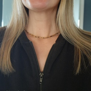 Puede incluir: Primer plano de una mujer con una sudadera con capucha negra con cremallera y un collar de cuentas de madera marrón. El collar es simple y delicado, con pequeñas cuentas redondas.