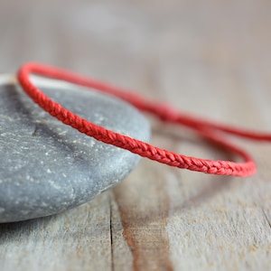 May include: A red braided cord bracelet, lying on a grey stone and a wooden surface.