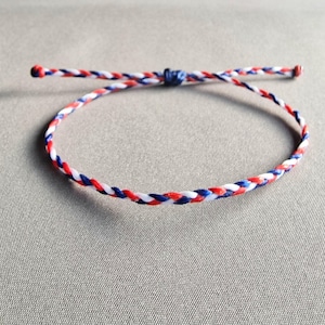 May include: Braided bracelet in red, white, and blue. The ends are knotted, and it has a sliding knot closure. The bracelet is adjustable and suitable for daily wear. The bracelet's design is simple and elegant.