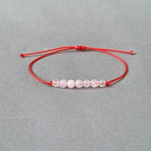 Könnte beinhalten: Ein rotes Armband mit rosa Perlen. Das Armband ist mit einem Knoten gebunden.