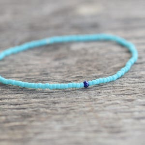 Op de afbeelding: Een delicate turquoise kralenarmband met een kleine, donkerblauwe accentkraal. De armband is gemaakt van kleine, ronde kralen en wordt gepresenteerd op een verweerd houten oppervlak.