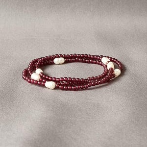 May include: A layered necklace with small, deep red beads and larger, white, oval pearls. The necklace is displayed on a grey surface, highlighting its elegant design and colour contrast.