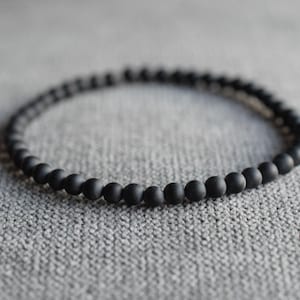 Könnte beinhalten: Ein kreisförmiges Armband, das aus kleinen, gleichmäßigen, mattschwarzen, kugelförmigen Perlen besteht. Die Perlen sind in einer durchgehenden Schleife angeordnet und bilden ein einfaches und konsistentes Design. Die Textur jeder Perle erscheint glatt und nicht reflektierend und bildet einen dunklen, festen Ring, der durchgehend eine einheitliche Größe aufweist.