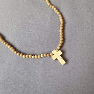 May include: A necklace featuring a series of small, round, light brown wooden beads strung together. A smooth, light-colored wooden cross pendant is attached to the center of the bead strand. The text Azul Sereno Jewelry is visible at the bottom.
