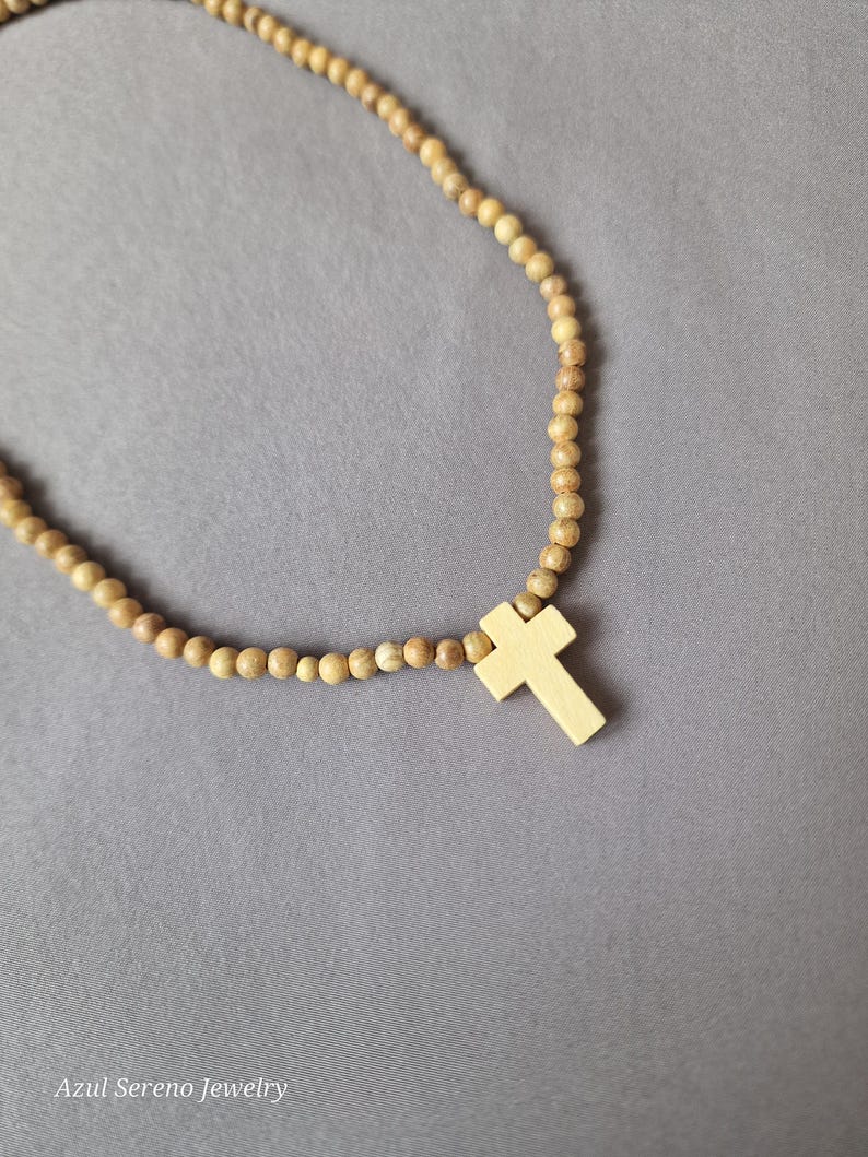 May include: A necklace featuring a series of small, round, light brown wooden beads strung together. A smooth, light-colored wooden cross pendant is attached to the center of the bead strand. The text Azul Sereno Jewelry is visible at the bottom.