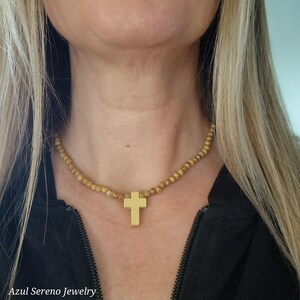 May include: A necklace featuring a series of small, round, light brown wooden beads strung together. A smooth, solid gold-colored cross pendant hangs from the center of the bead strand. The necklace rests against the neck, showcasing the contrast between the natural wood texture and the metallic finish of the cross.